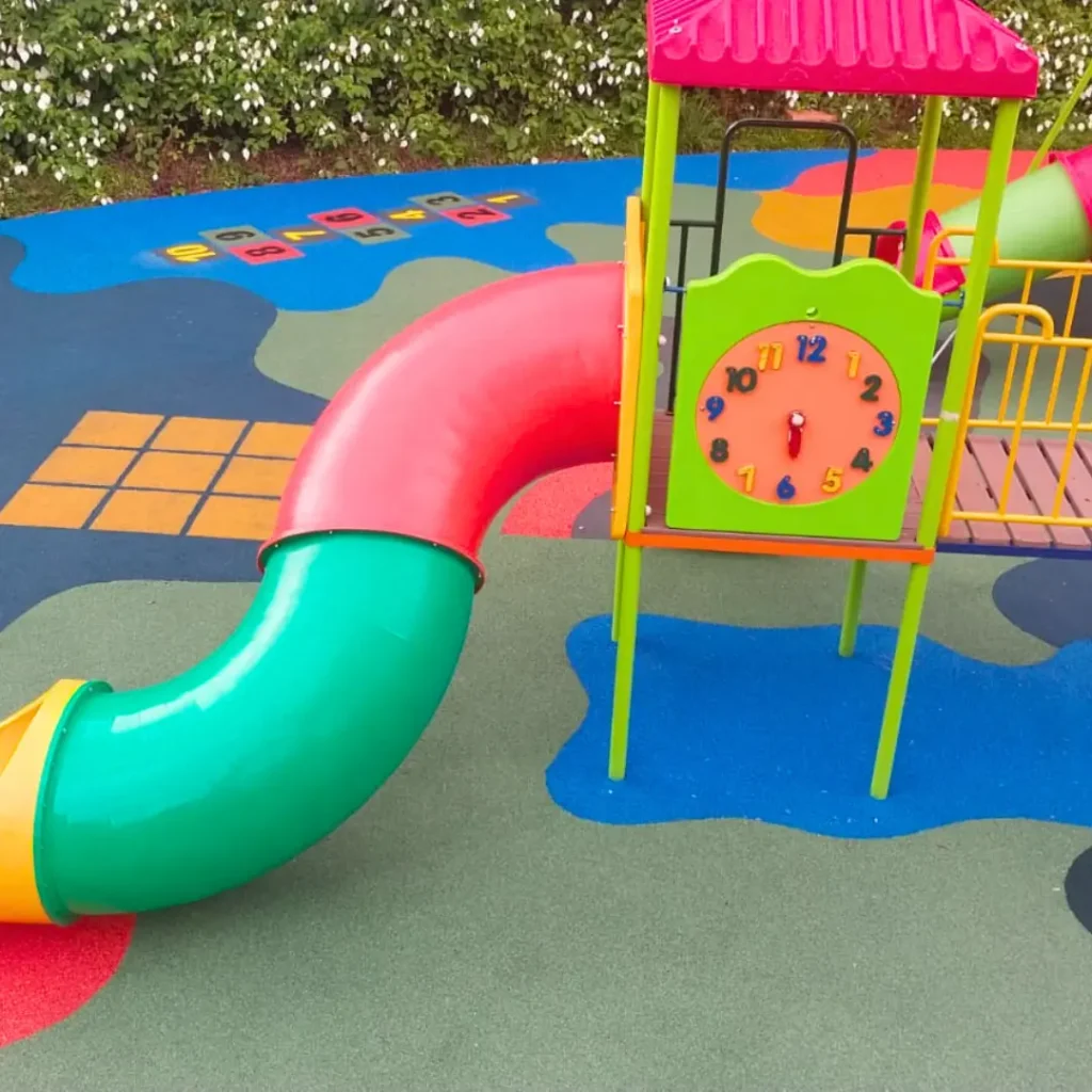 Um parque infantil com piso contínuo na paginação de cores Amarela, Azul, Rosa Salmão com um escorregador, balanços e uma estrutura de brincar.