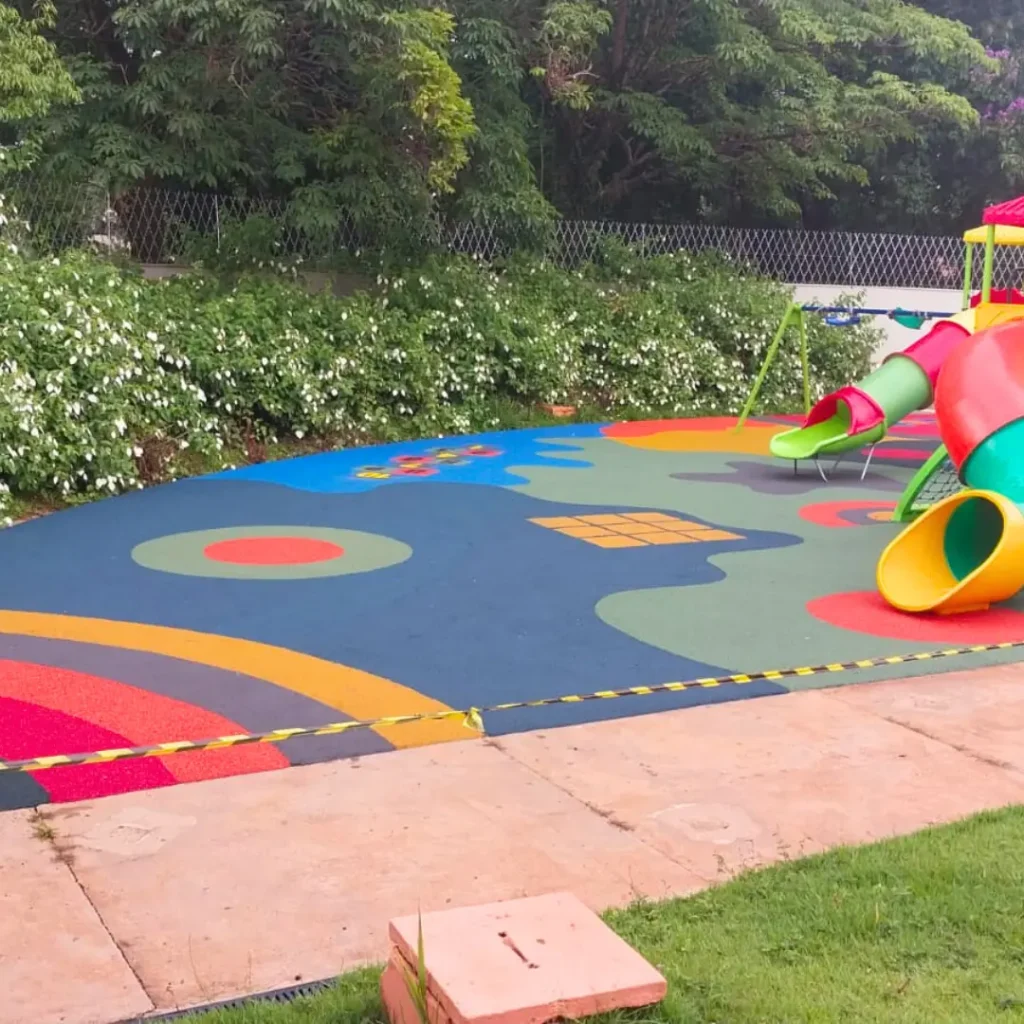 Um parque infantil com piso contínuo na paginação de cores Amarela, Azul, Rosa Salmão com um escorregador, balanços e uma estrutura de brincar.