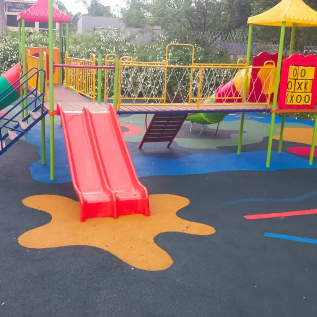 Um parque infantil com piso contínuo na paginação de cores Amarela, Azul, Rosa Salmão com um escorregador, balanços e uma estrutura de brincar.