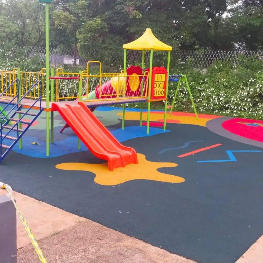 Um parque infantil com piso contínuo na paginação de cores Amarela, Azul, Rosa Salmão com um escorregador, balanços e uma estrutura de brincar.