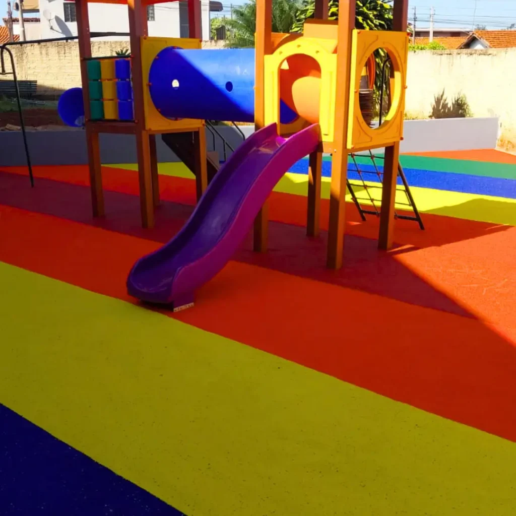 Um parque infantil com piso contínuo na paginação de cores Amarela, Azul, Rosa Salmão com um escorregador, balanços e uma estrutura de brincar.