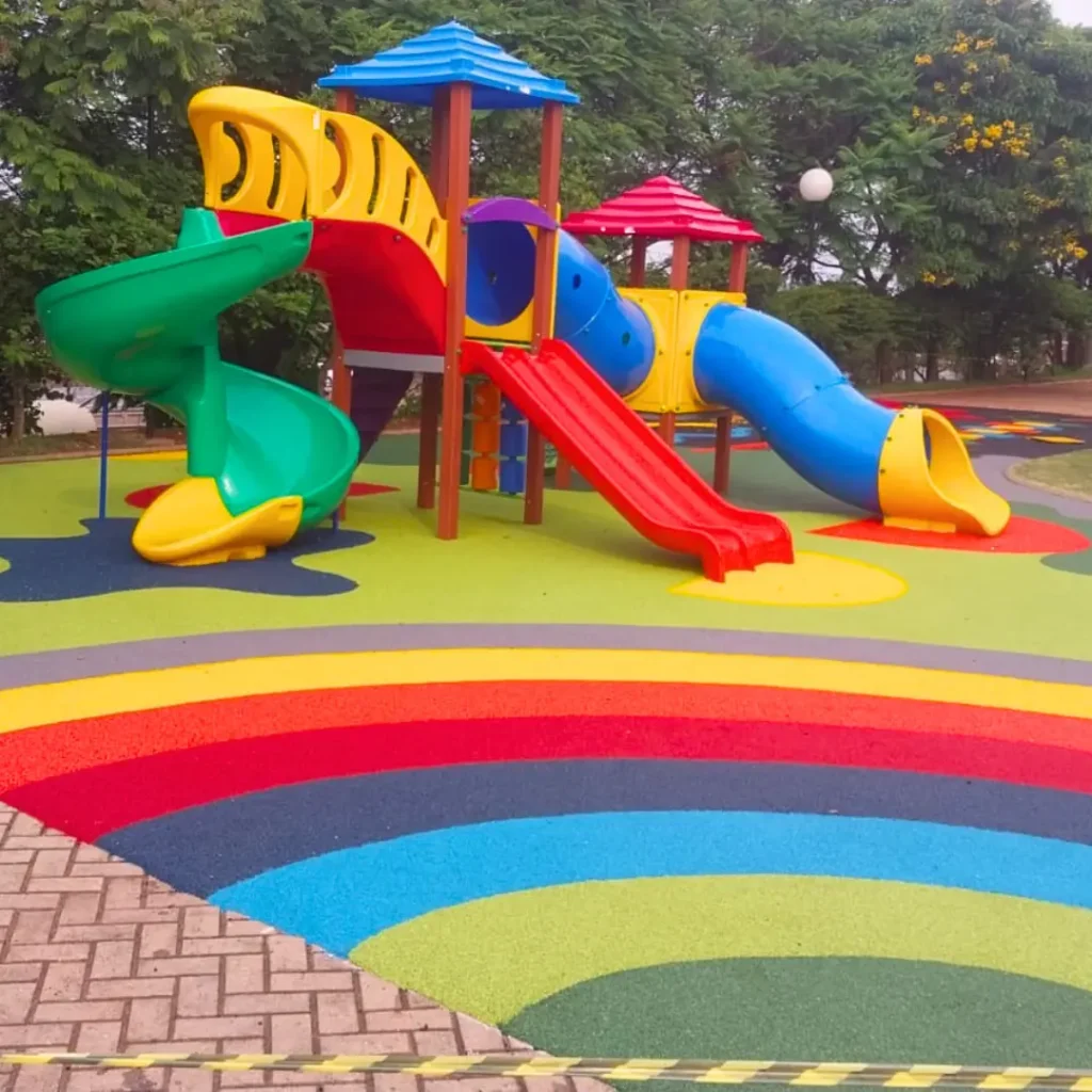 Um parque infantil com piso contínuo na paginação de cores Amarela, Azul, Rosa Salmão com um escorregador, balanços e uma estrutura de brincar.