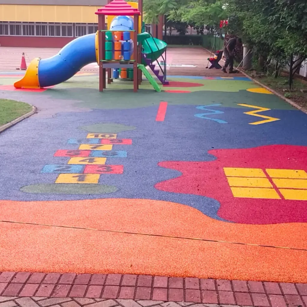 Um parque infantil com piso contínuo na paginação de cores Amarela, Azul, Rosa Salmão com um escorregador, balanços e uma estrutura de brincar.